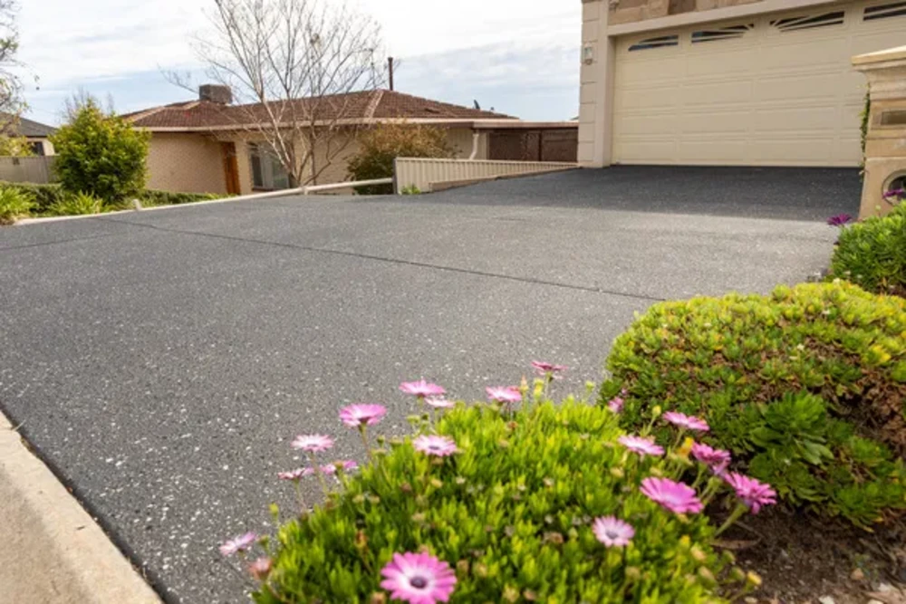 Dark speckled concrete driveway in front of a home with pink flowers in the foreground
