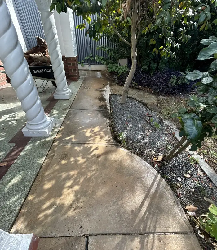 Worn, stained concrete path beside white twisted veranda posts and a garden, before resurfacing