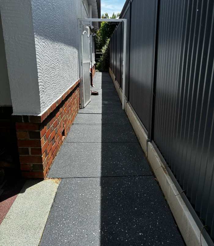 Narrow dark grey resurfaced concrete side path between a rendered wall and a metal fence