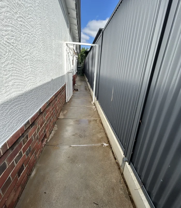 Narrow concrete side path between a rendered wall and a dark metal fence, before resurfacing