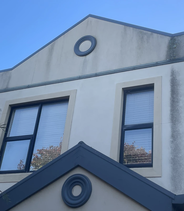 Rendered house facade with a round window, stained and dull under a blue sky, before pressure washing