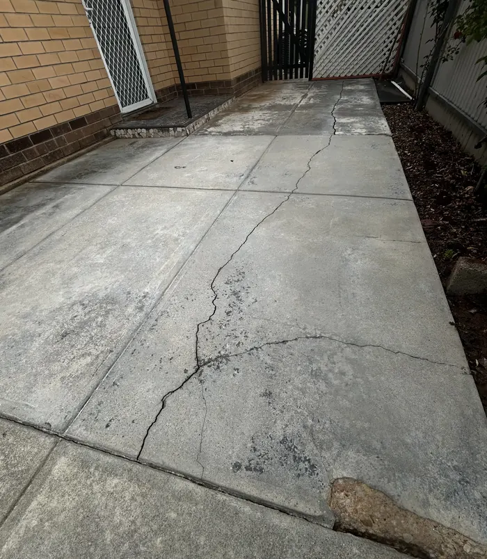 Cracked, stained concrete side path beside a brick wall and fence, before concrete resurfacing