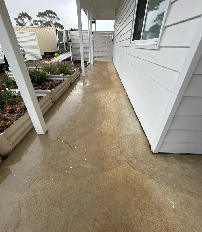 Stained, golden-brown concrete veranda floor beside a white weatherboard wall, before epoxy flooring