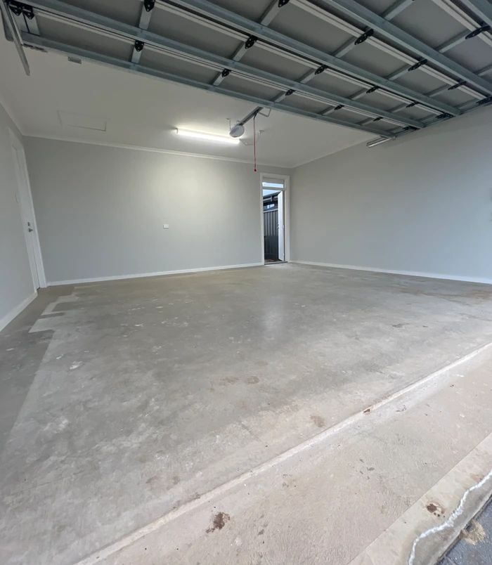 Bare concrete garage floor with white walls and a roller door, before epoxy flooring