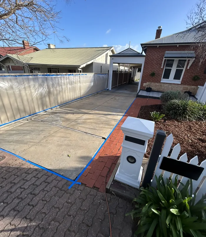 Pale concrete driveway with blue masking tape beside a timber fence and brick home, before resurfacing