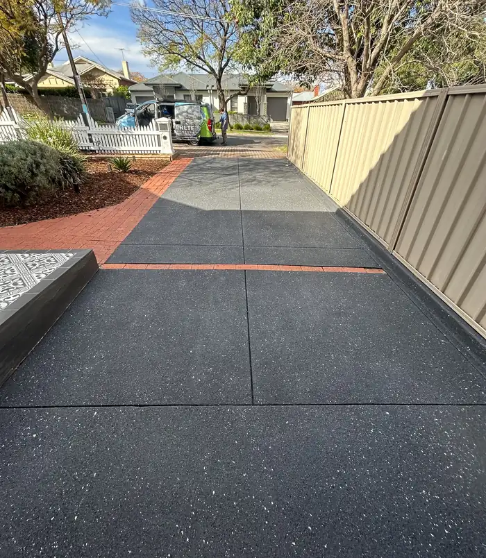 Dark grey resurfaced concrete driveway with red brick edging beside a metal fence