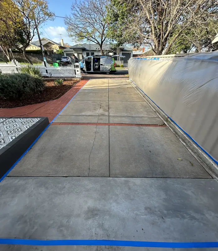 Pale concrete driveway with blue masking tape along a metal fence and brick edging, before resurfacing