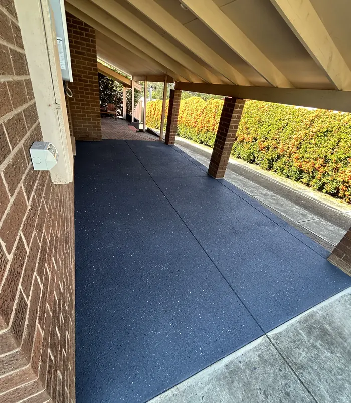 Dark resurfaced concrete floor under a carport beside a brick wall and orange hedge