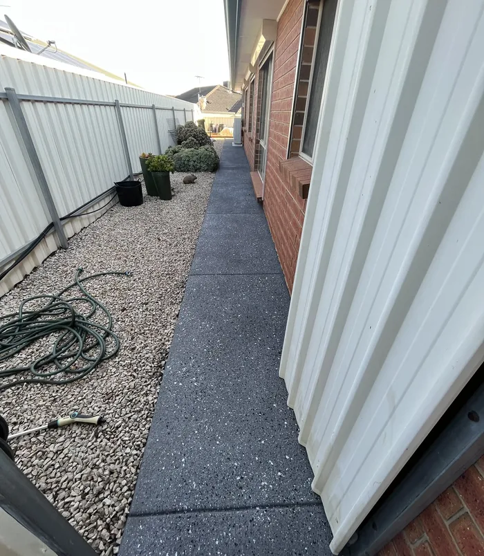 Dark resurfaced concrete side path beside a brick wall, white fence and gravel garden