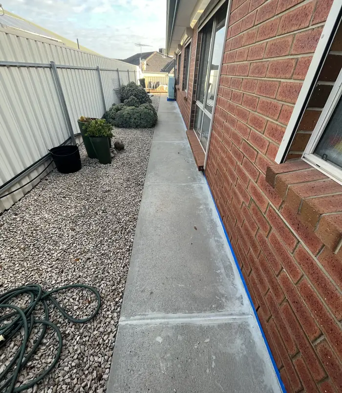 Pale, worn concrete side path beside a brick wall, white fence and gravel garden, before resurfacing