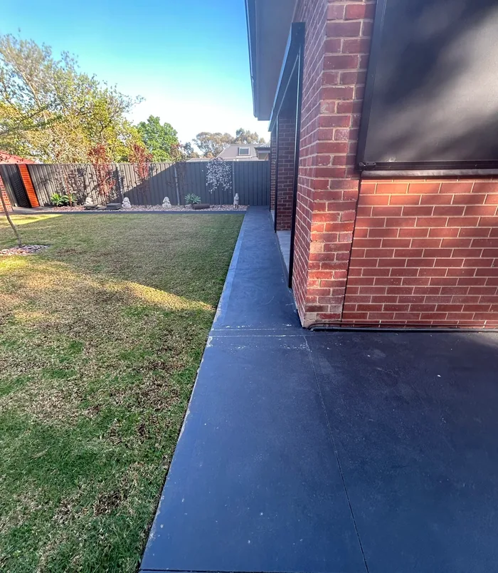 Narrow concrete path beside a red brick wall and lawn with a blue coating, before resurfacing is finished