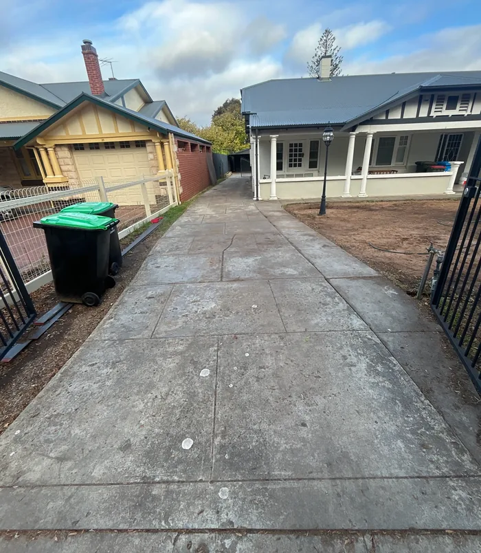 Cracked, patchy concrete driveway beside a fence and green wheelie bin, before concrete resurfacing