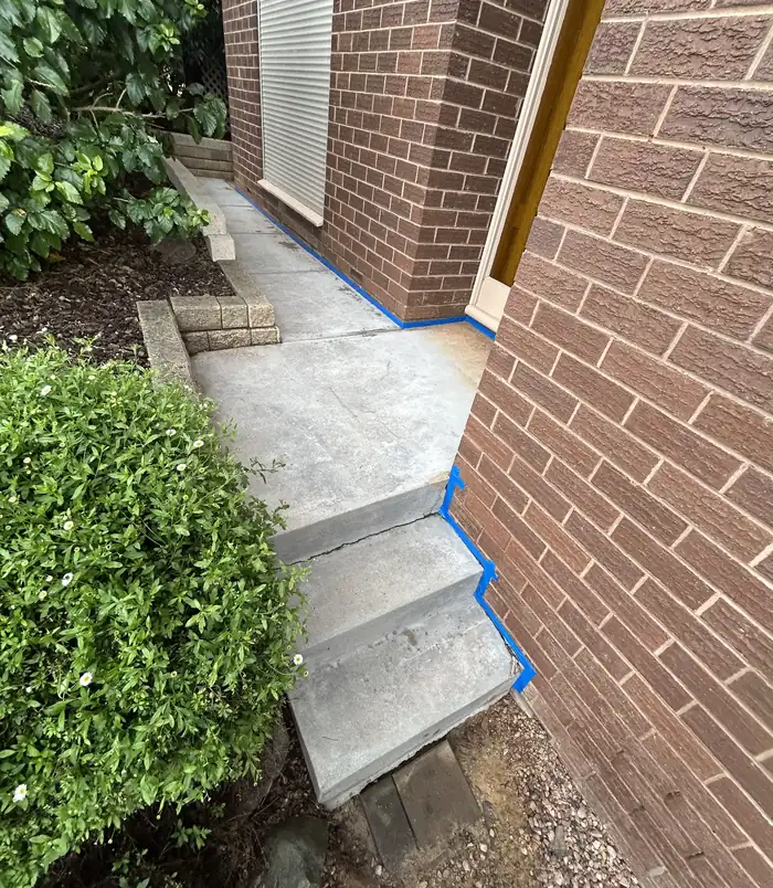 Rough concrete path beside a brick wall with blue masking tape along the edge, before resurfacing