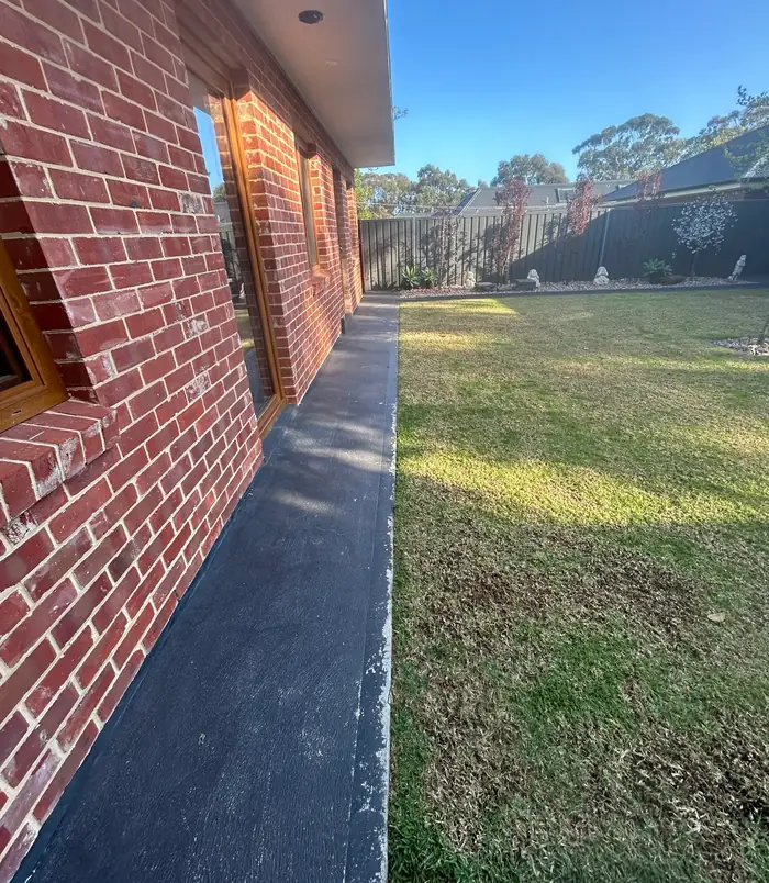 Narrow concrete path beside a red brick wall and lawn, before concrete resurfacing