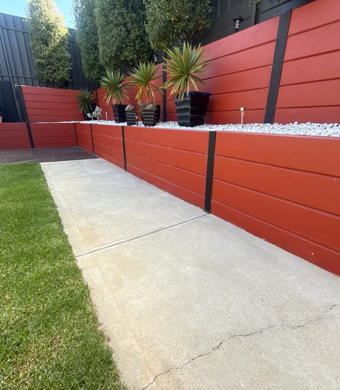 Pale, stained concrete path beside a red retaining wall and lawn, before concrete resurfacing