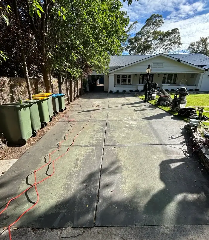 Long, patchy concrete driveway beside green wheelie bins and a cream home, before concrete resurfacing