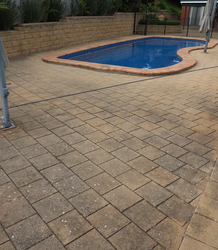 Dull, dirty brick pavers around a freeform blue swimming pool, before pressure washing