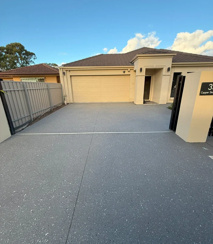 Freshly resurfaced dark grey concrete driveway in front of a home with a tan garage door