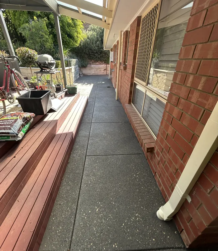 Dark grey resurfaced concrete side path beside a brick wall and timber deck