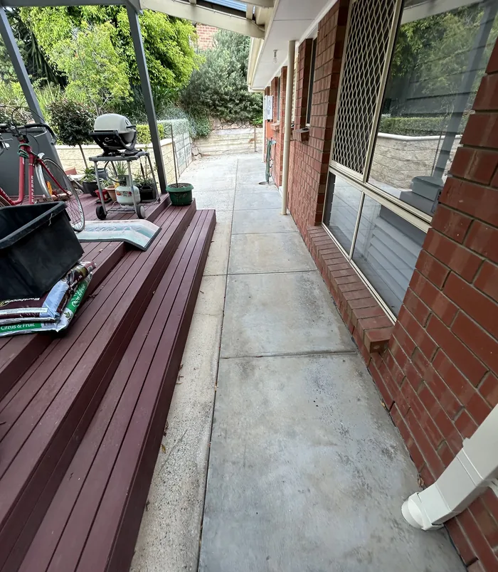 Pale concrete side path beside a brick wall, timber deck and garden, before resurfacing