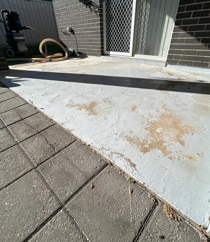 Stained, rust-marked concrete patio beside a dark brick wall and paving, before epoxy flooring