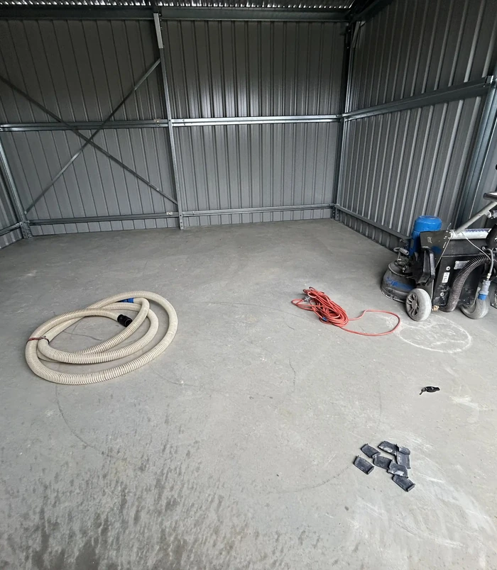 Bare concrete floor in a large metal shed with floor grinding equipment and a hose, before epoxy flooring