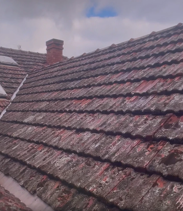 Dull, faded red tiled roof with a chimney under a cloudy sky, before pressure washing