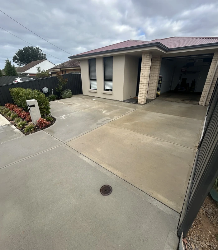 Pale, weathered concrete driveway in front of a home with a red roof and garden bed, before resurfacing