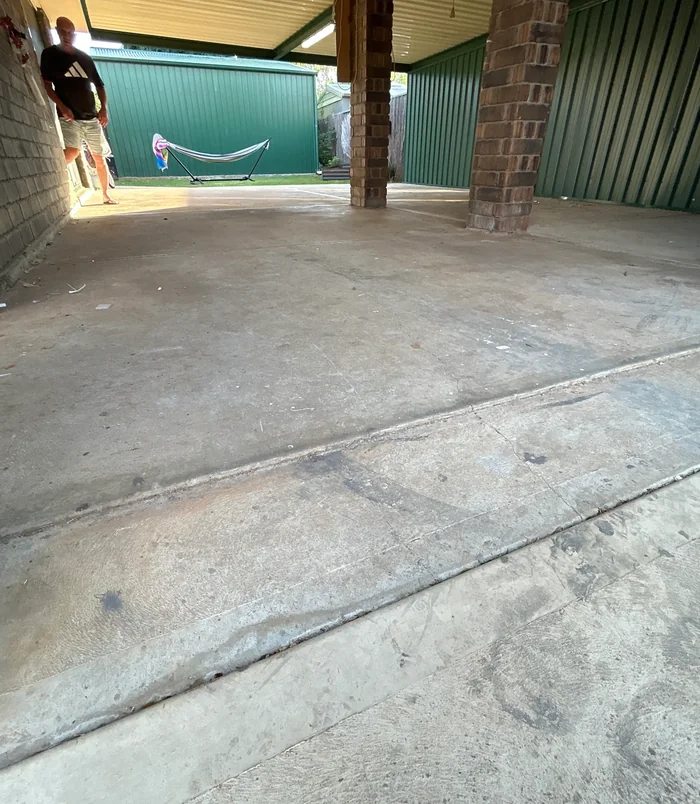 Stained, grimy concrete carport floor beside brick pillars and a green shed, before epoxy flooring