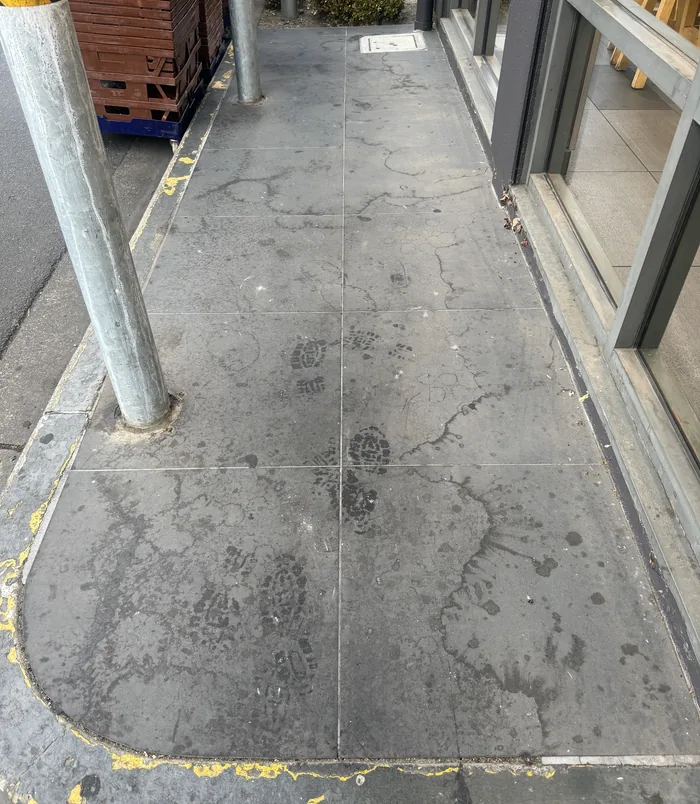Dull, stained concrete walkway beside a shopfront with steel posts, before pressure washing