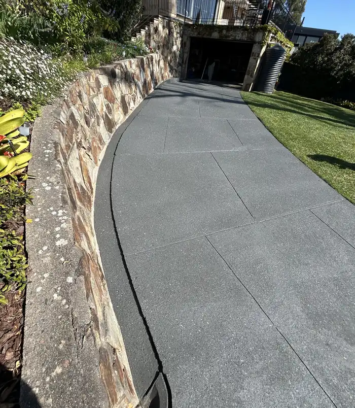 Dark grey resurfaced concrete path curving beside a stone retaining wall and lawn