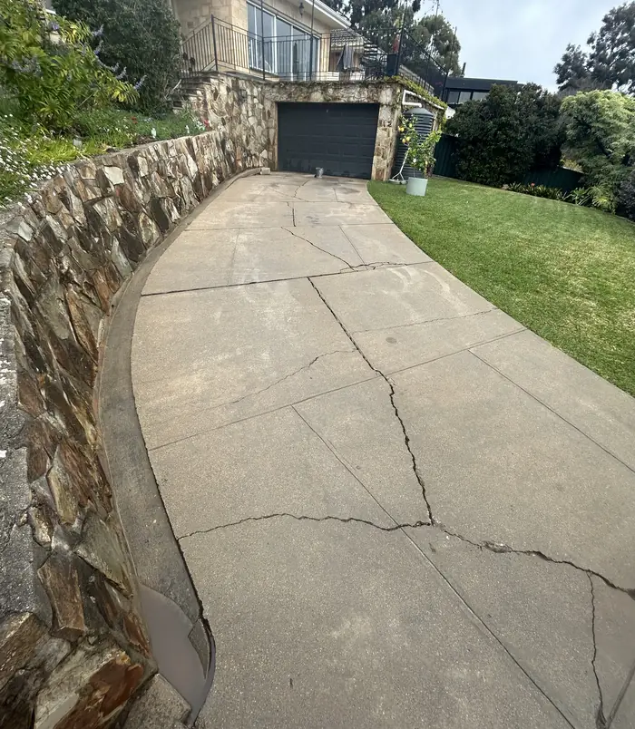 Pale, cracked concrete path curving beside a stone retaining wall and lawn, before resurfacing