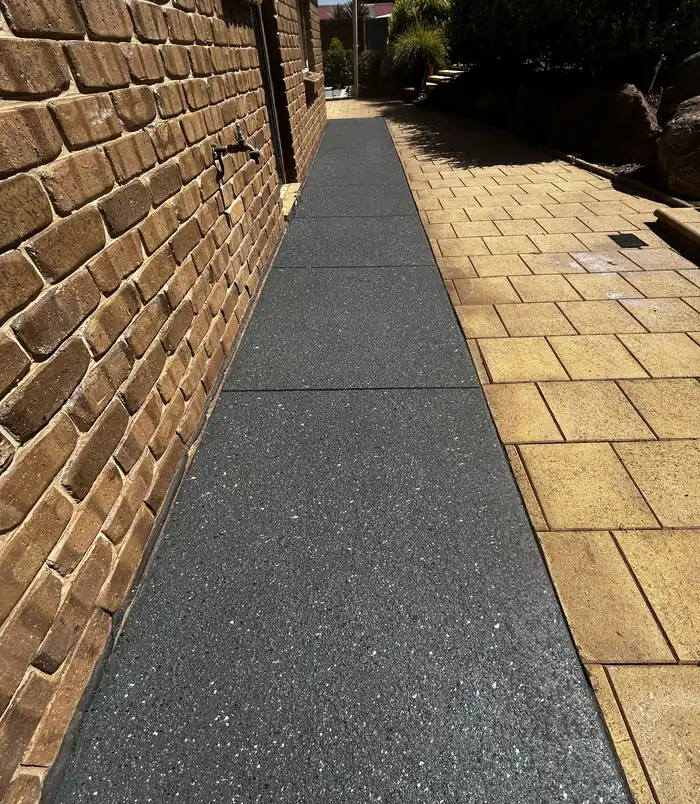 Dark grey resurfaced concrete path between golden pavers and a brick wall