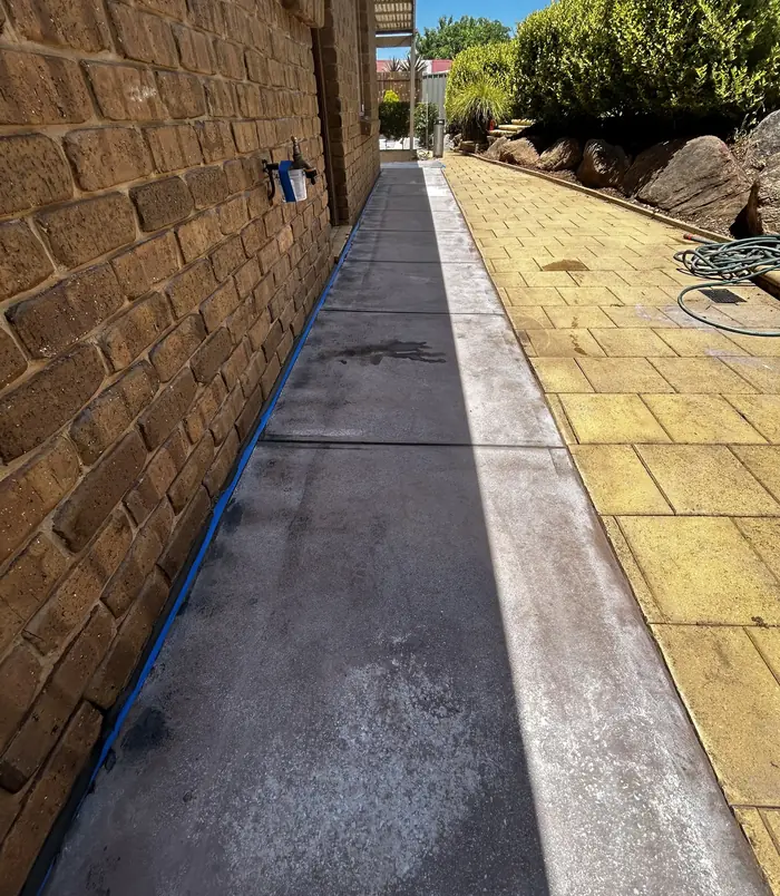 Narrow concrete strip with blue masking tape between golden pavers and a brick wall, before resurfacing