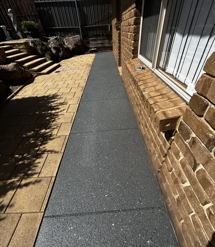Dark speckled resurfaced concrete path between golden pavers and a brick wall