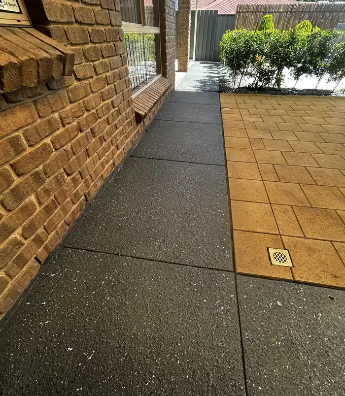 Speckled dark resurfaced concrete path between a brick wall and golden pavers
