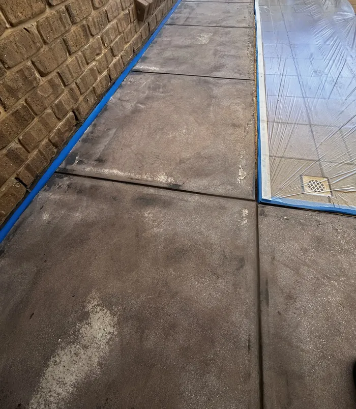 Stained concrete path with blue masking tape beside a brick wall and paving, before resurfacing