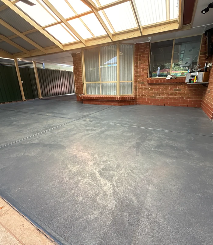 Stained concrete patio floor under a pergola beside a brick wall, before epoxy flooring