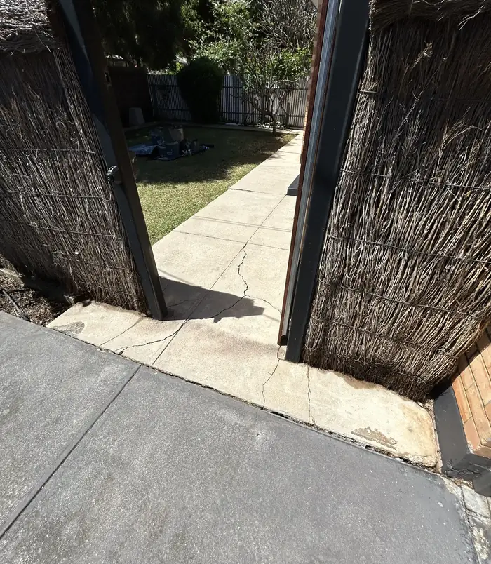 Pale concrete side path between a woven brush fence and a dark gate, before resurfacing