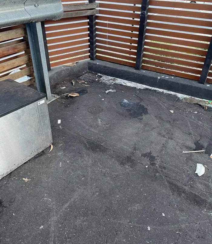 Dirty, debris-covered concrete floor in an enclosed area with timber slat screens, before pressure washing
