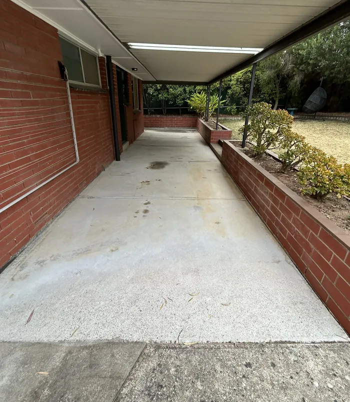 Pale, worn concrete veranda floor beside a brick wall and garden bed, before epoxy flooring