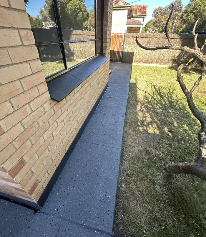 Dark grey resurfaced concrete path beside a brick wall, window ledge and lawn