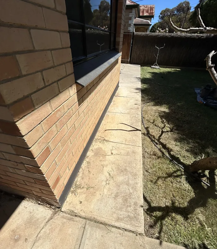 Pale, weathered concrete path beside a brick wall and lawn, before concrete resurfacing