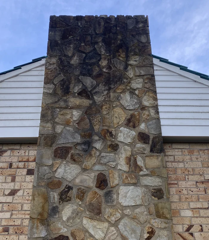 Dark, stained stone chimney on a weatherboard house under a blue sky, before pressure washing