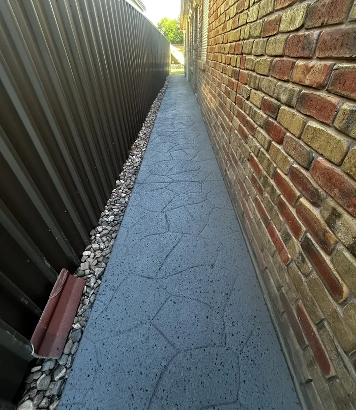 Narrow dark grey resurfaced stamped concrete side path beside a brick wall with gravel edging