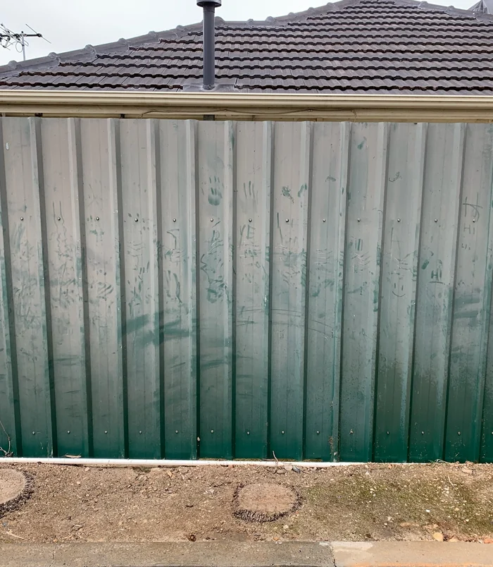 Green metal fence with white streaks and dirt marks beneath a tiled roof, before pressure washing