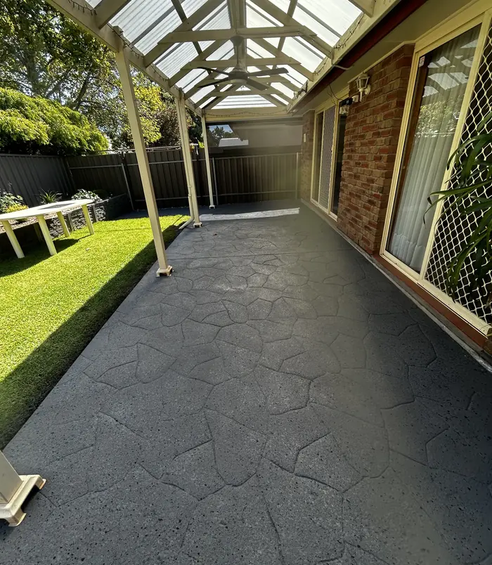 Dark grey resurfaced concrete walkway under a pergola beside a brick home and lawn