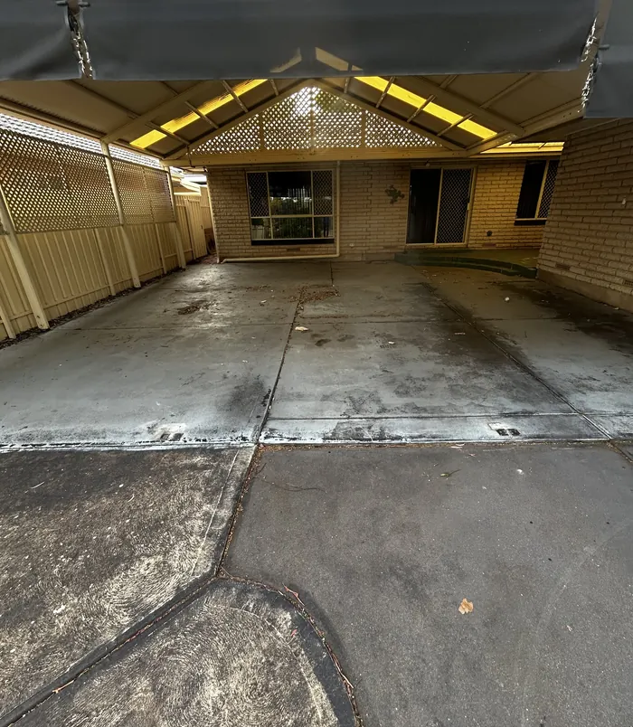 Dirty, stained concrete floor under a pergola beside a brick home, before concrete resurfacing