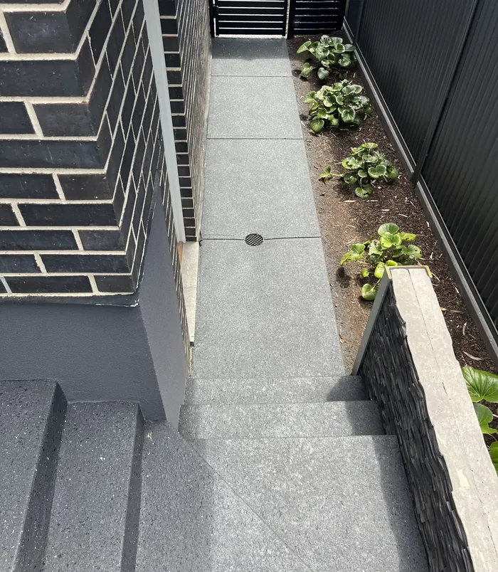 Smooth speckled grey resurfaced concrete side path beside a dark brick wall and garden bed