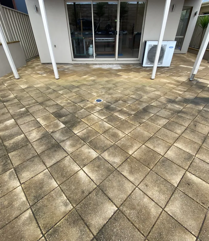 Dirty, algae-stained pavers under a pergola beside sliding glass doors, before pressure washing